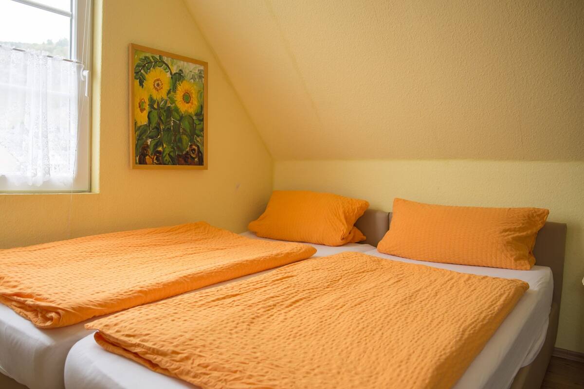Quarto com duas camas, cada uma com colcha e almofadas laranja, um quadro de girassóis na parede e uma janela com cortina translúcida.