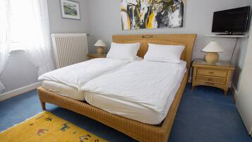 Un lit double avec du linge de lit blanc, une tête de lit en bois, deux tables de chevet avec des lampes, un téléviseur posé sur un meuble et un tableau au-dessus du lit.