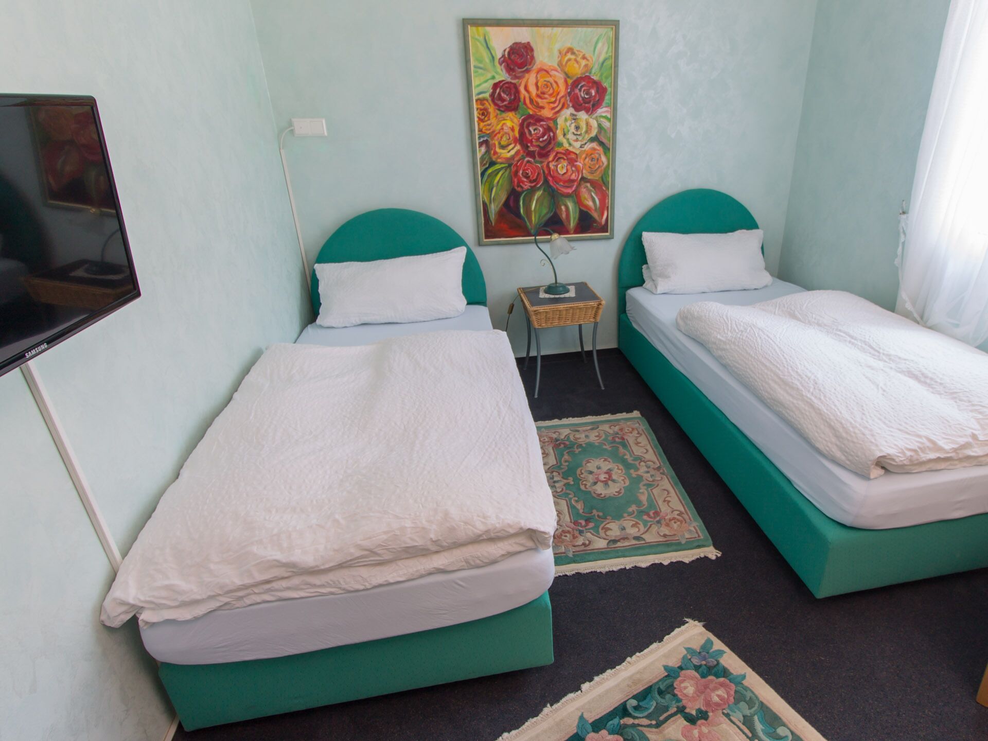 Una habitación con dos camas individuales, una mesita pequeña, un televisor montado en la pared y un cuadro con motivos florales.