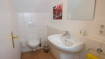 A bathroom with a white sink, a toilet, a trash bin, and a yellow towel on a rack.