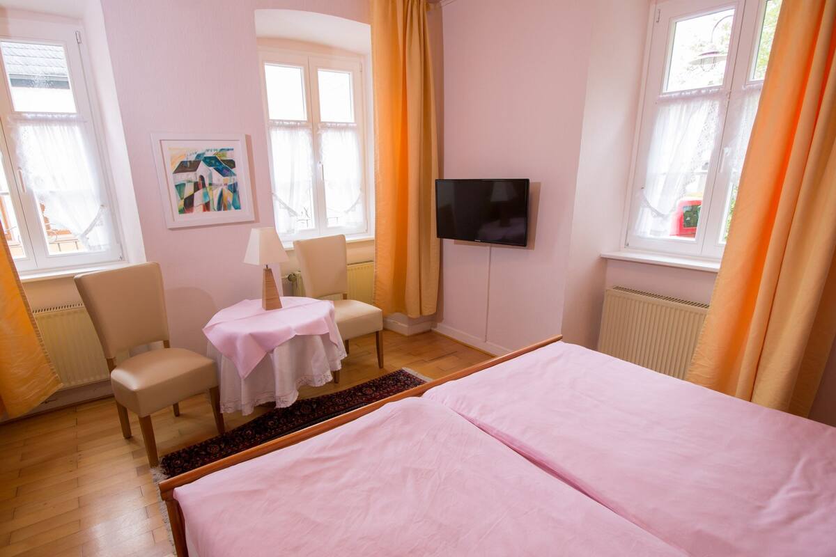 Quarto com cama, escrivaninha, cadeira, televisão e janela com cortinas.