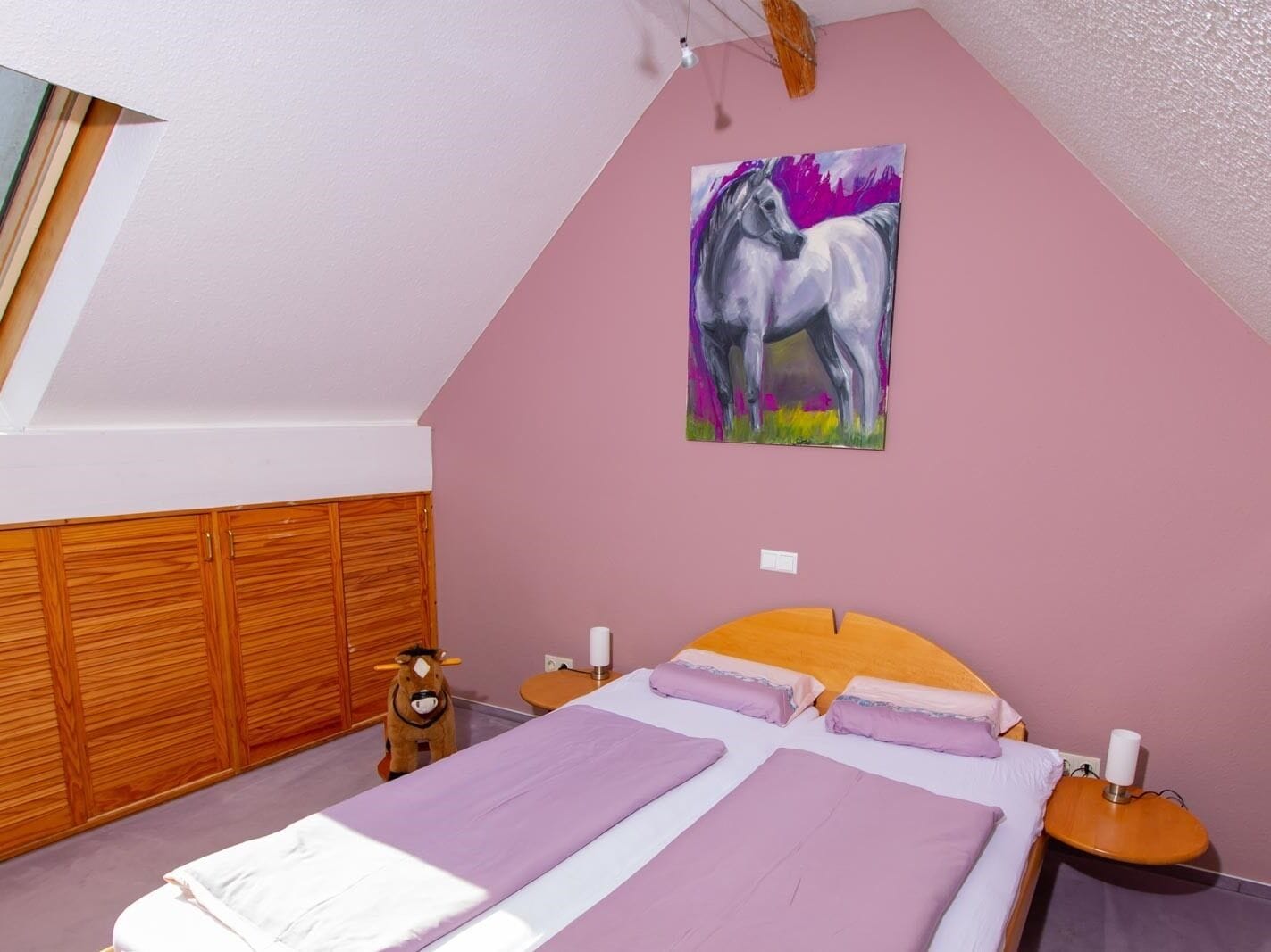 A bedroom with a sloped ceiling, a bed with purple bedding, a wooden wardrobe, a small wooden nightstand, a wall-mounted painting of a horse, and a window with a wooden blind.