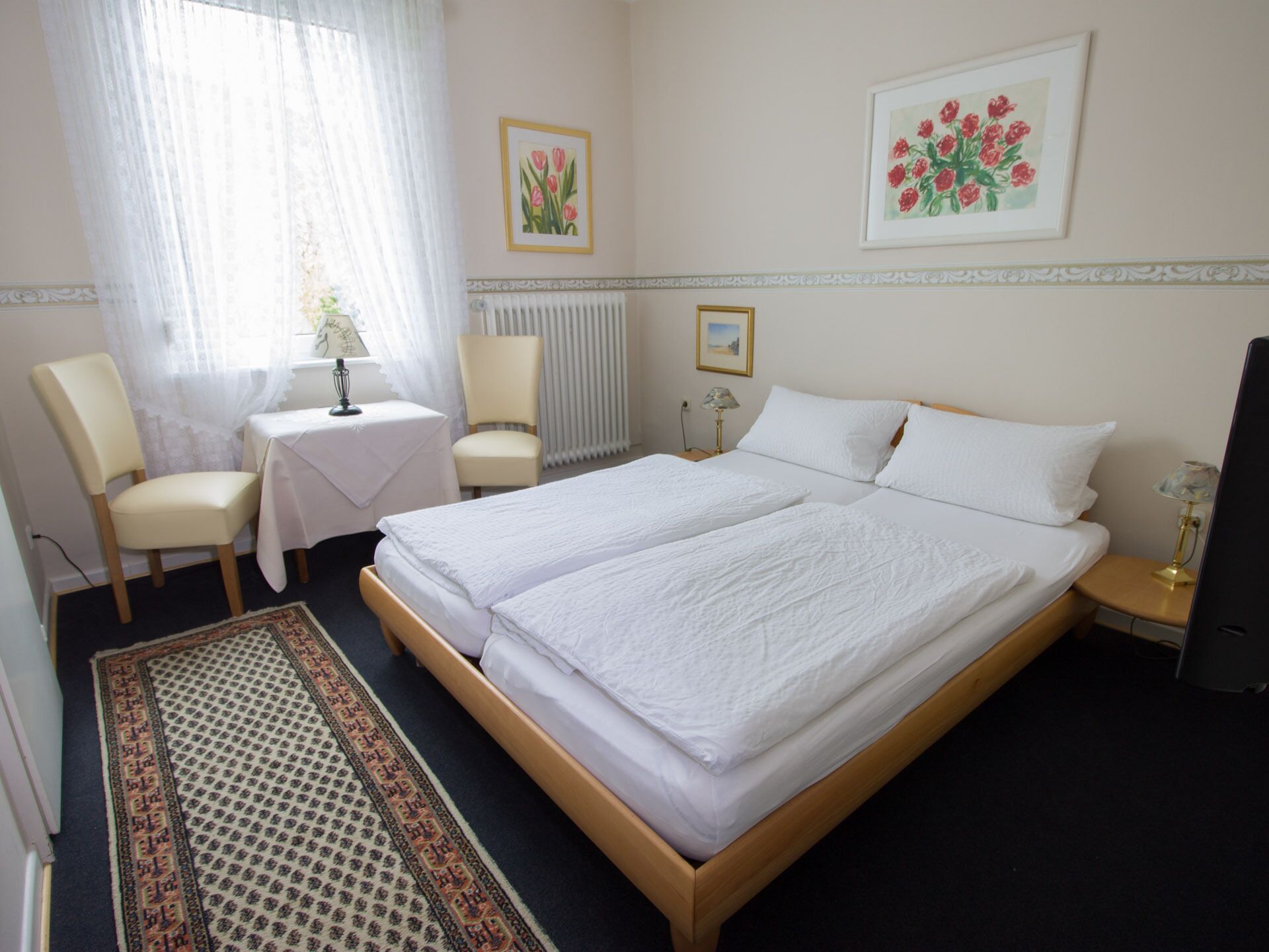 A hotel room with two single beds, a small table, a chair, a radiator, and two framed pictures on the wall.