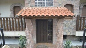 Een gebouw met een pannendak, stenen muren en een houten deur. Er staan planten in potten op de grond.