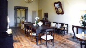 Ein Esszimmer mit einem dunklen Holztisch, weißen Stühlen und einer Bank. Eine Chandelüster, ein Spiegel an der Wand und eine Tür mit dekorativem Rahmen sind ebenfalls vorhanden.