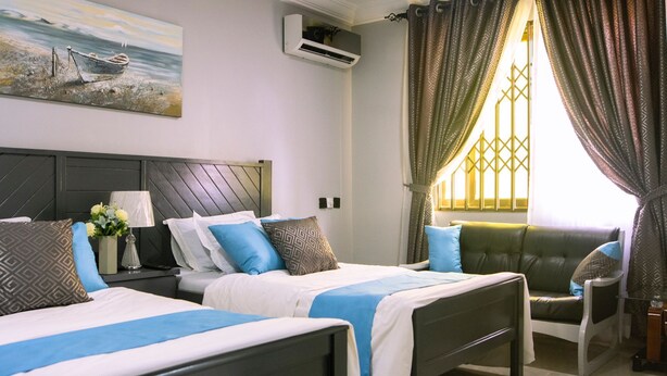 A hotel room with two beds, a green sofa, a decorative window with patterned curtains, and a wall-mounted air conditioner. - KCS Guest House (Accra)