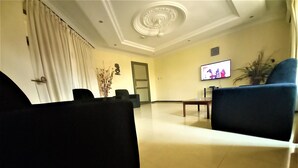 A spacious living room with a flat-screen TV mounted on the wall, a coffee table, and a blue armchair. - KCS Guest House (Accra)