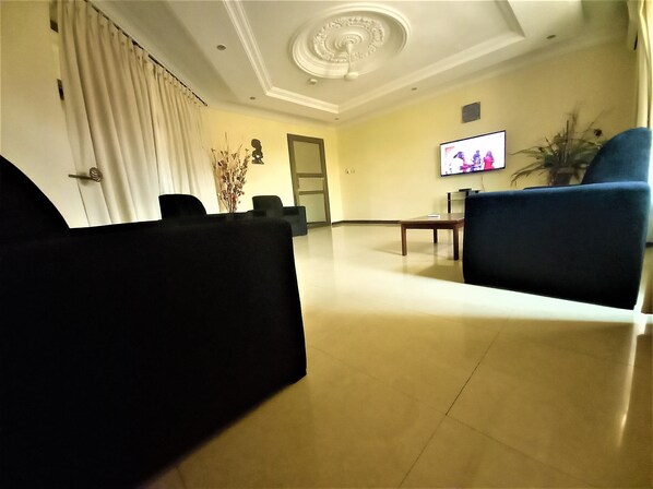 A spacious living room with a flat-screen TV mounted on the wall, a coffee table, and a blue armchair. - KCS Guest House (Accra)