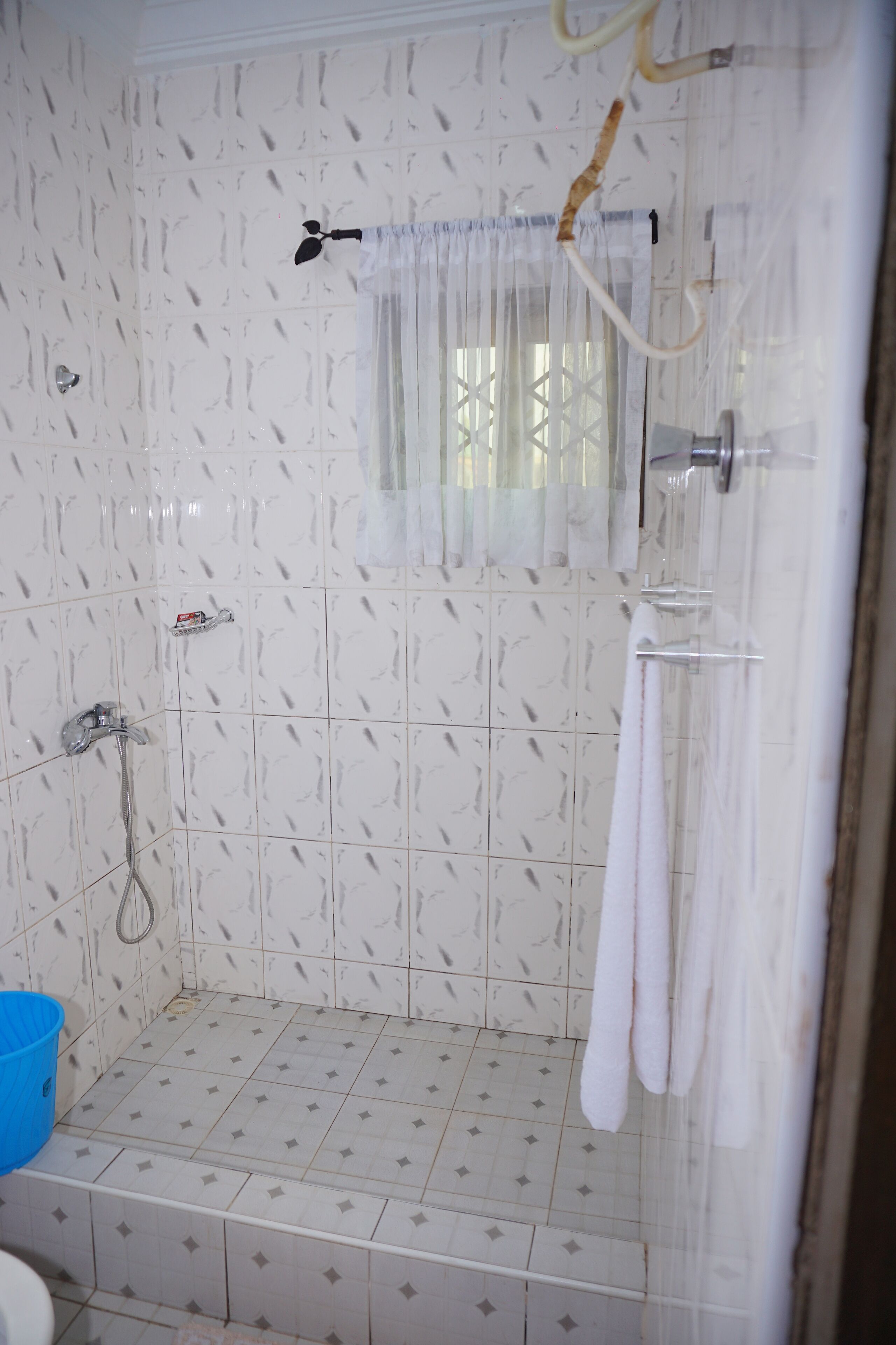 Un baño con ducha, paredes de azulejos, una ventana con cortina y un cubo azul.