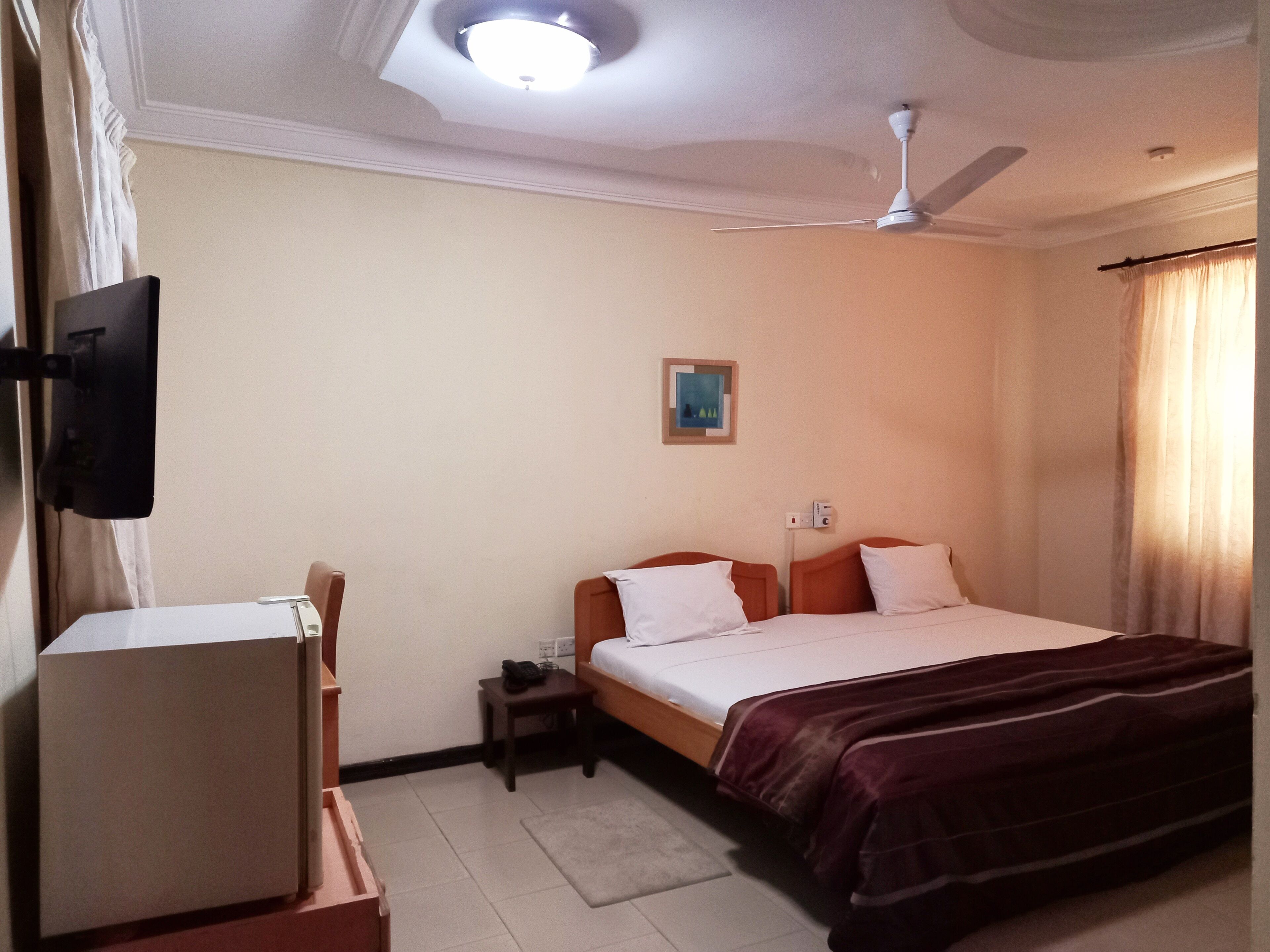A hotel room with a bed, a desk, a chair, a ceiling fan, and a painting on the wall.