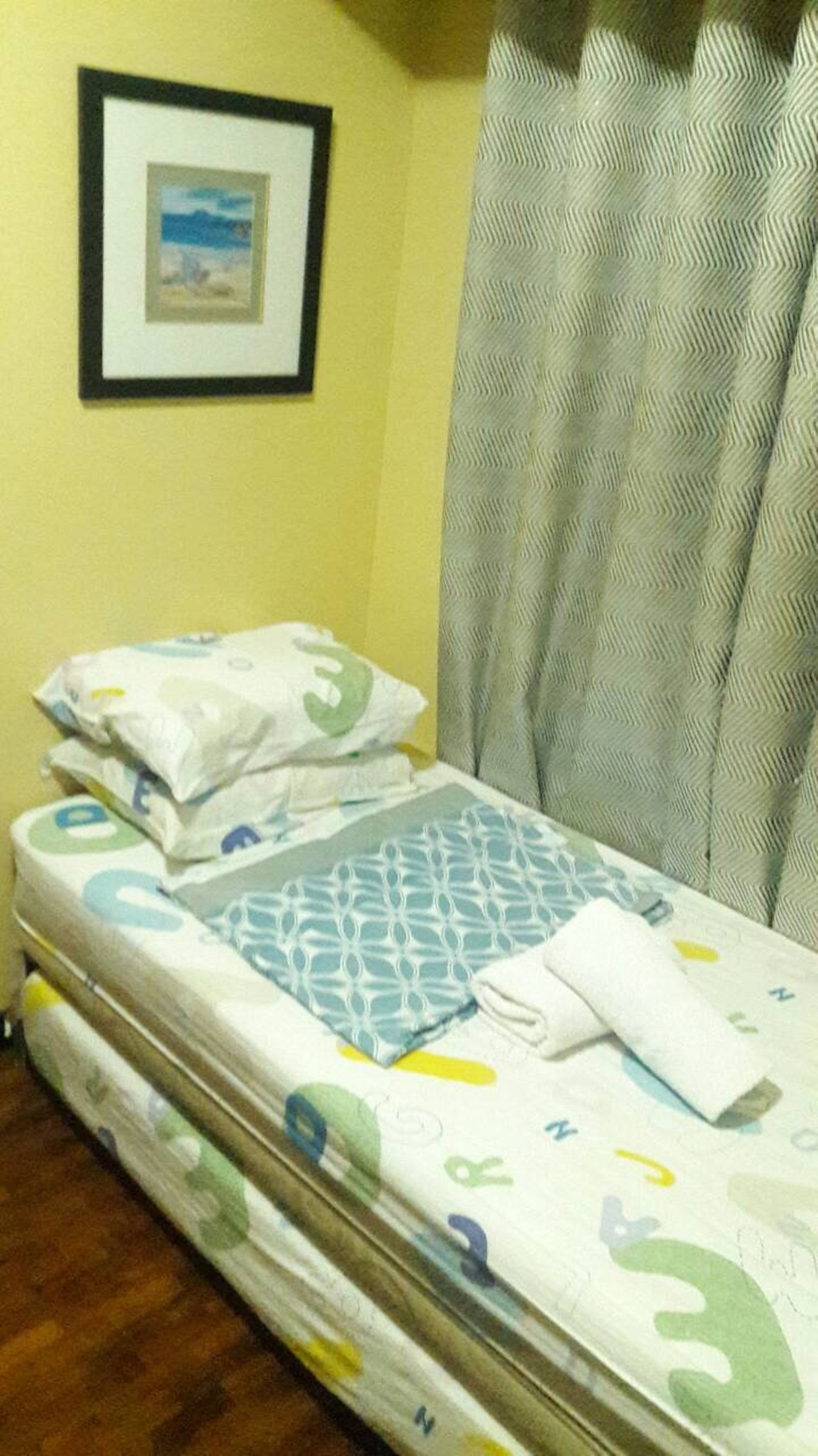 Una cama bien hecha con una colcha estampada, almohadas y una toalla doblada. Un cuadro enmarco cuelga de la pared sobre la cama.