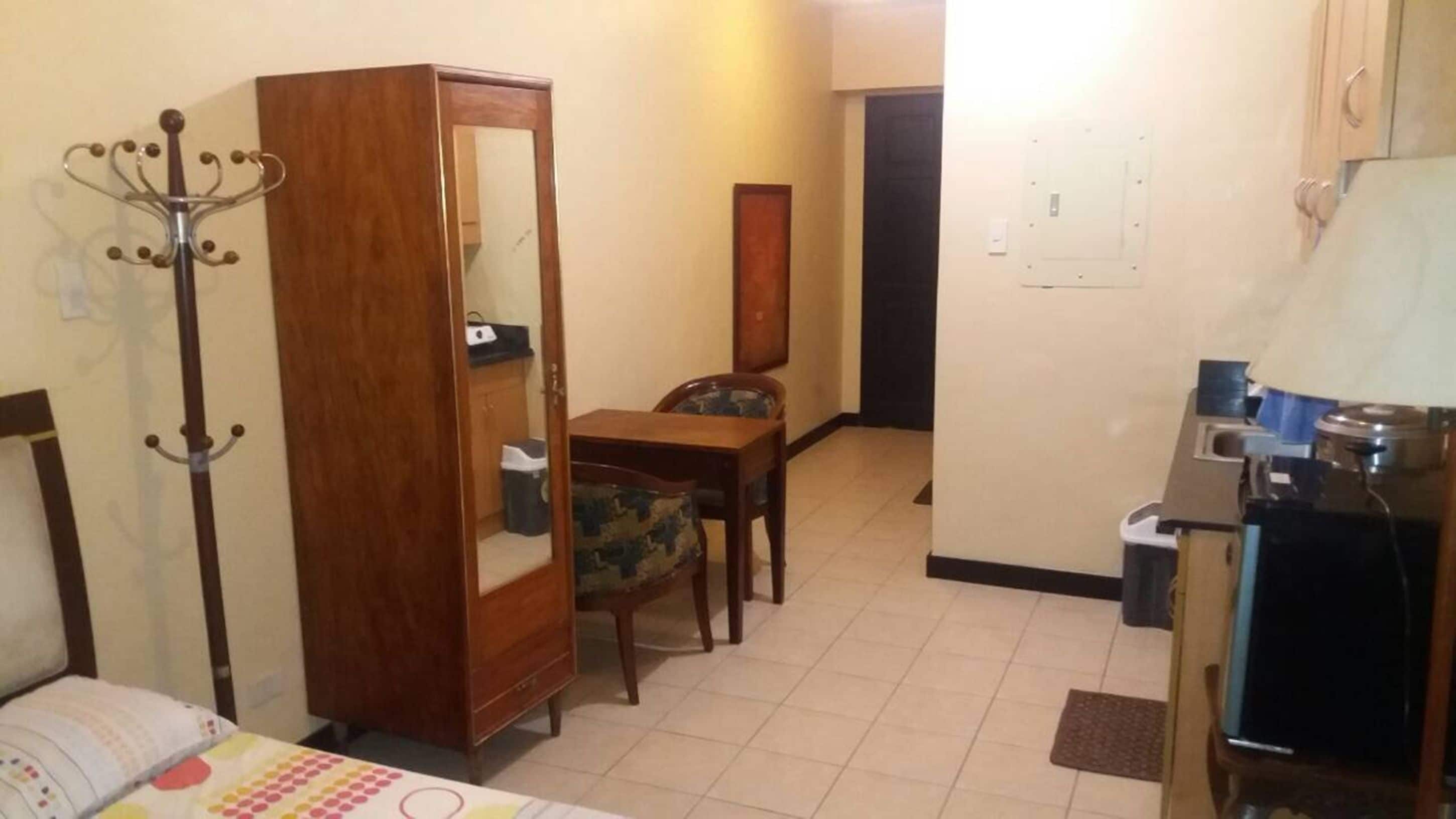 A hotel room with a bed, a wooden wardrobe, a chair, a small table, and a kitchenette area.