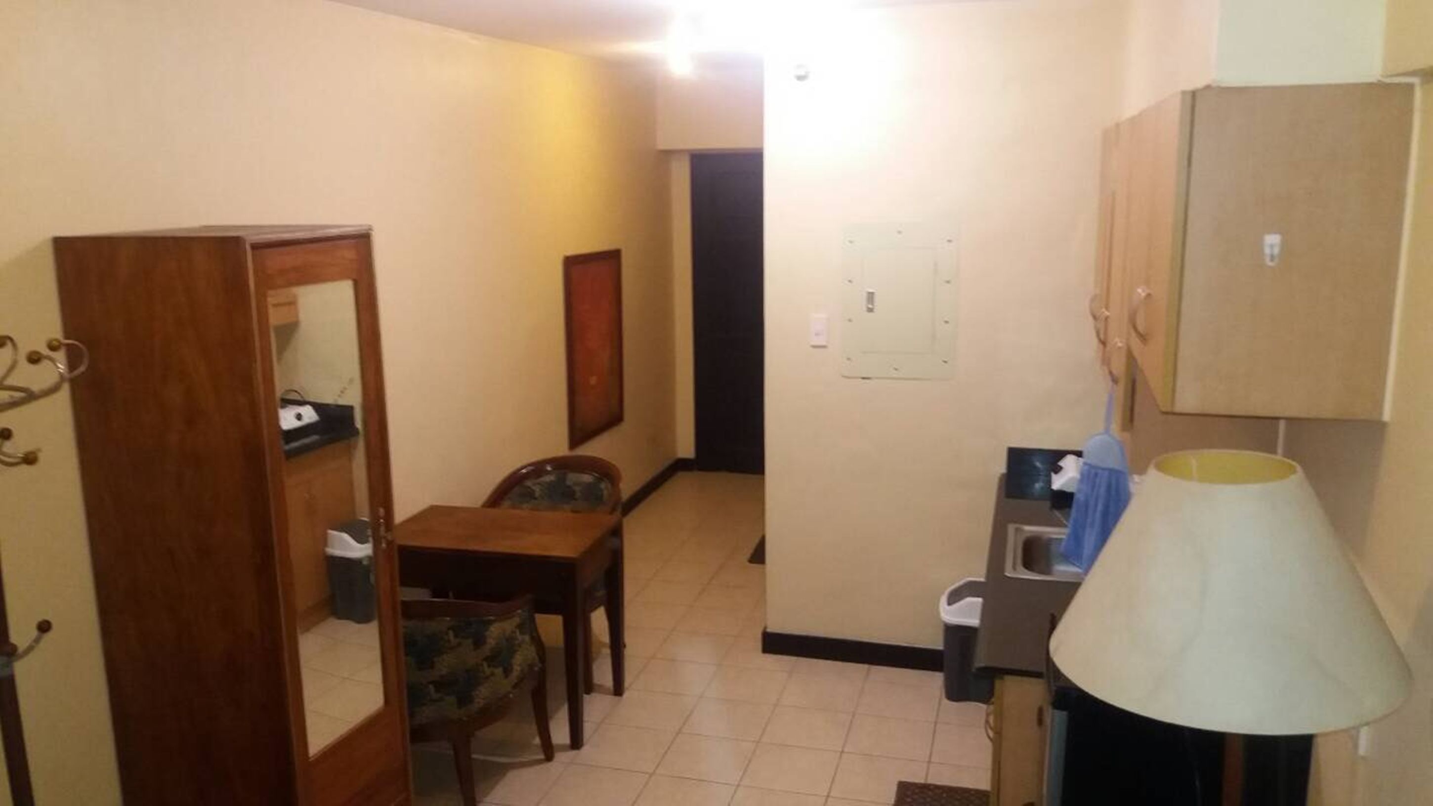 A compact room with a wooden dining table, a chair, a small kitchenette with a sink and a lamp, and a door leading to another room.