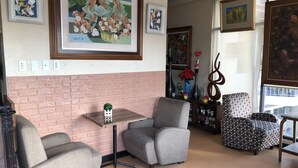 Un salon avec deux fauteuils gris, une petite table et une bibliothèque. Les murs sont ornés d’œuvres d’art encadrées et une grande fenêtre laisse entrer la lumière naturelle.