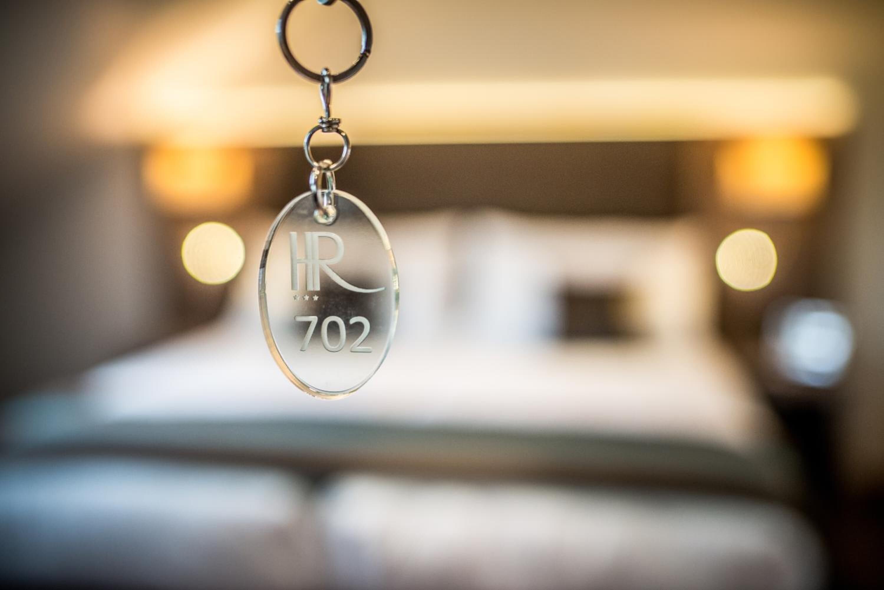 Un porte-clés avec la mention « IR 702 » gravée, suspendu devant une chambre d’hôtel floue, dans laquelle on aperçoit un lit et une table de chevet.