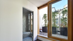 Une salle de bain moderne avec une douche à l’italienne en verre, un carrelage au sol et une grande fenêtre donnant sur un jardin et un immeuble.
