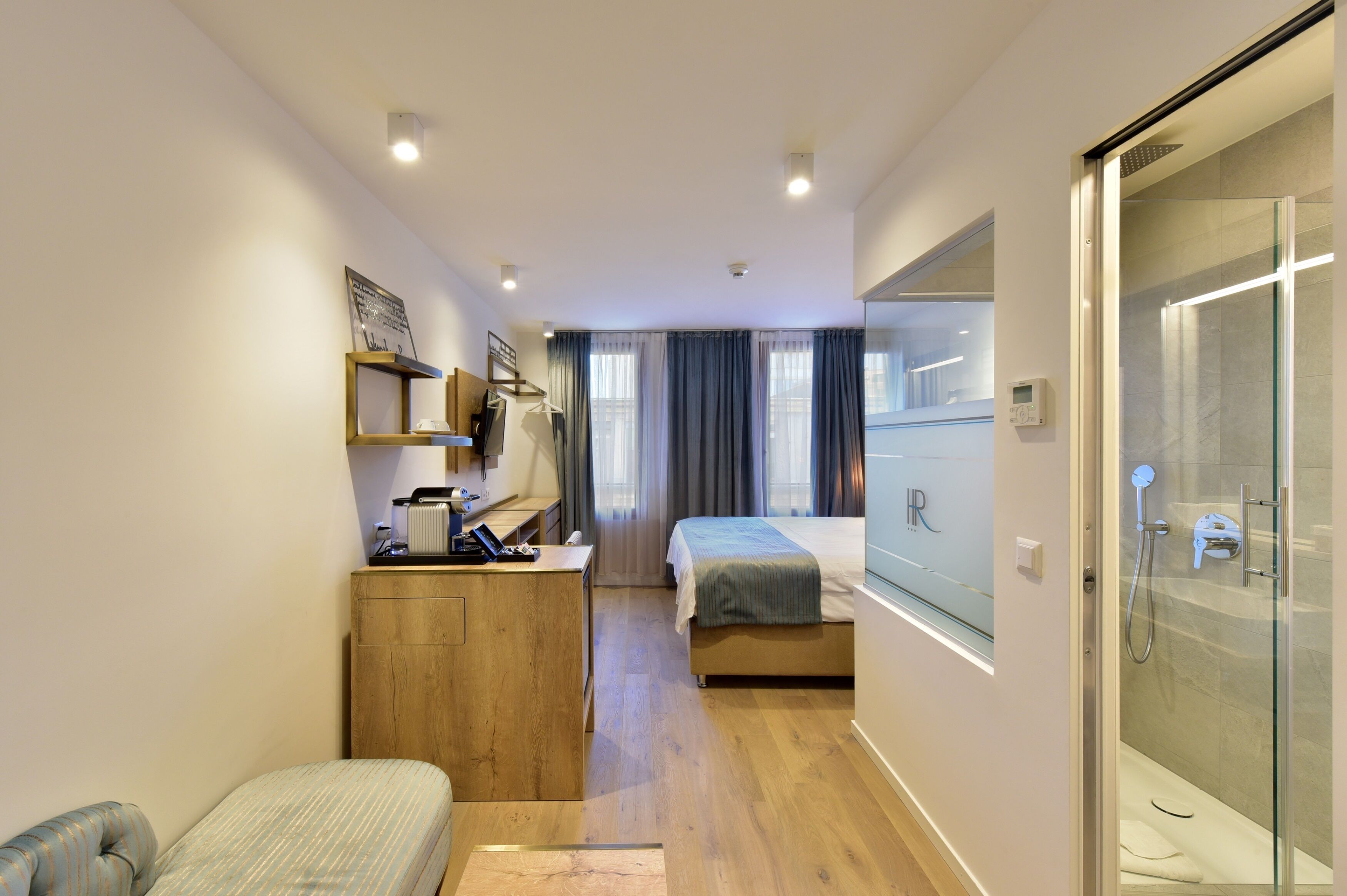 A compact hotel room with a bed, a desk, a chair, a small couch, a shower, and a large window with curtains.