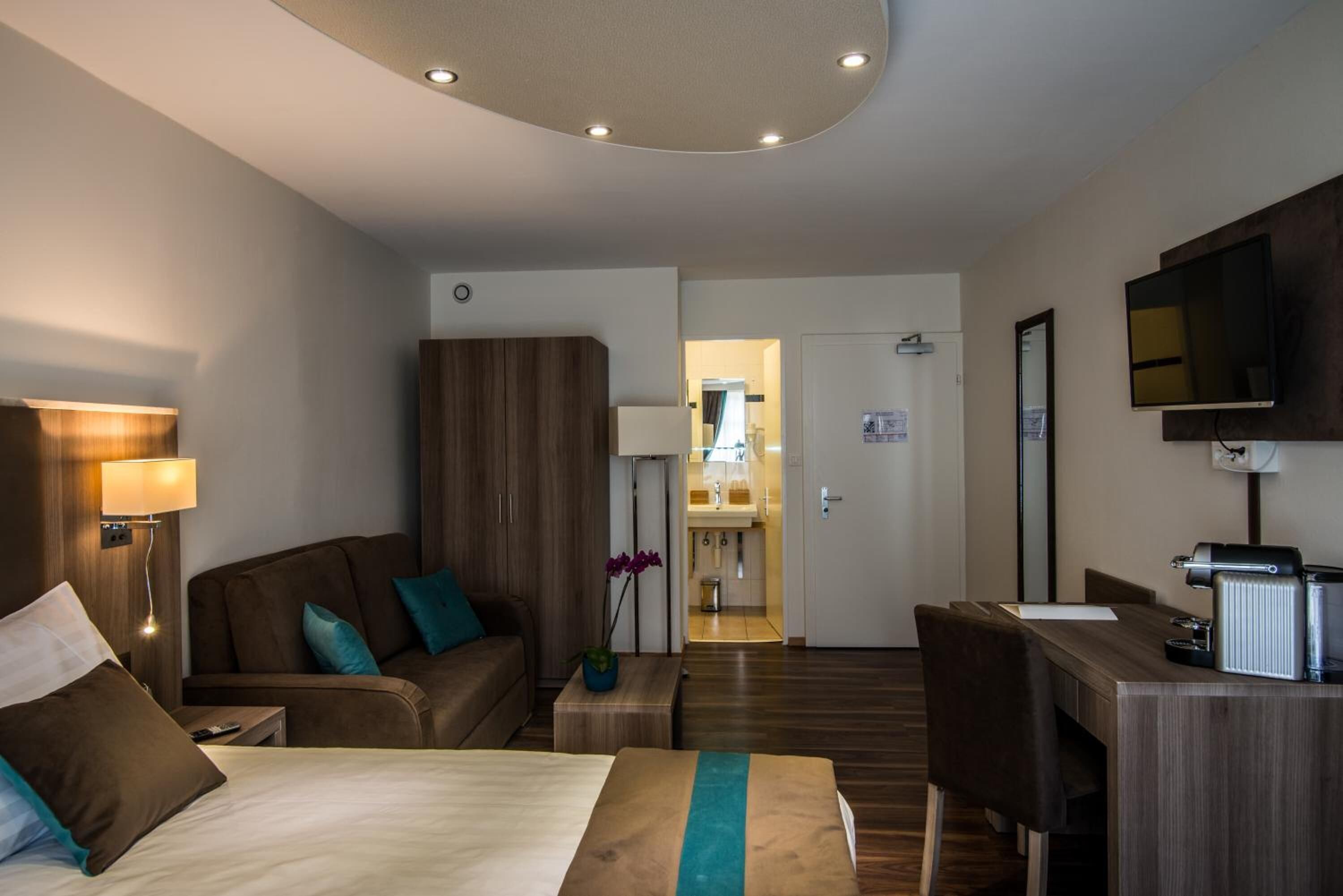 Ein Hotelzimmer mit Bett, Schreibtisch, Stuhl, Kleiderschrank, Fernseher und einem durch eine geöffnete Tür einsehbaren Badezimmer.