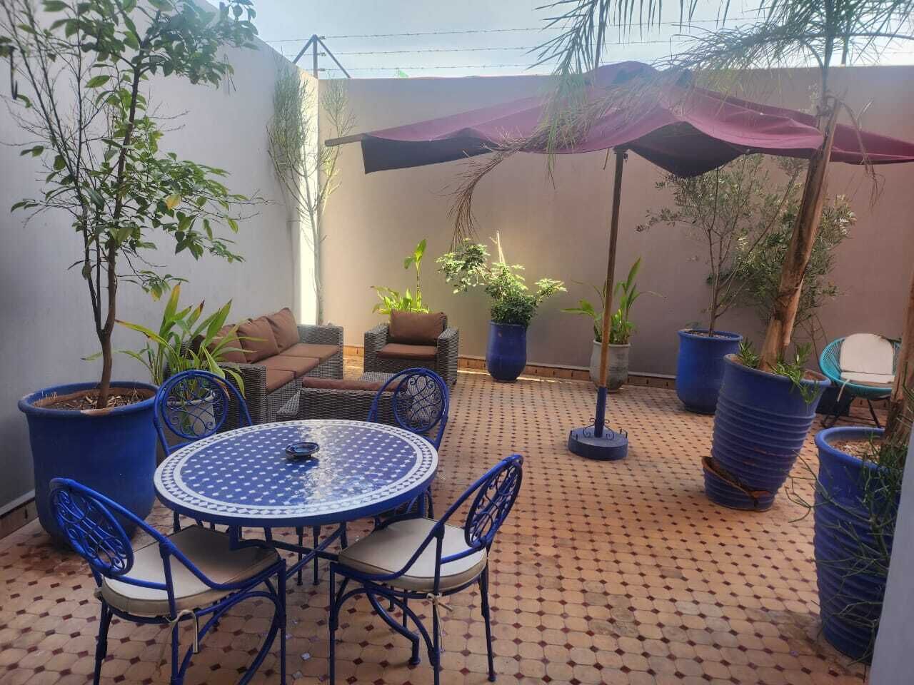 Un patio exterior con una mesa redonda y sillas, rodeado de macetas con plantas y un paraguas grande.