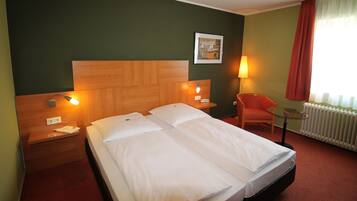 A hotel room with a double bed, two bedside tables, a chair, a small table, a lamp, and a radiator.