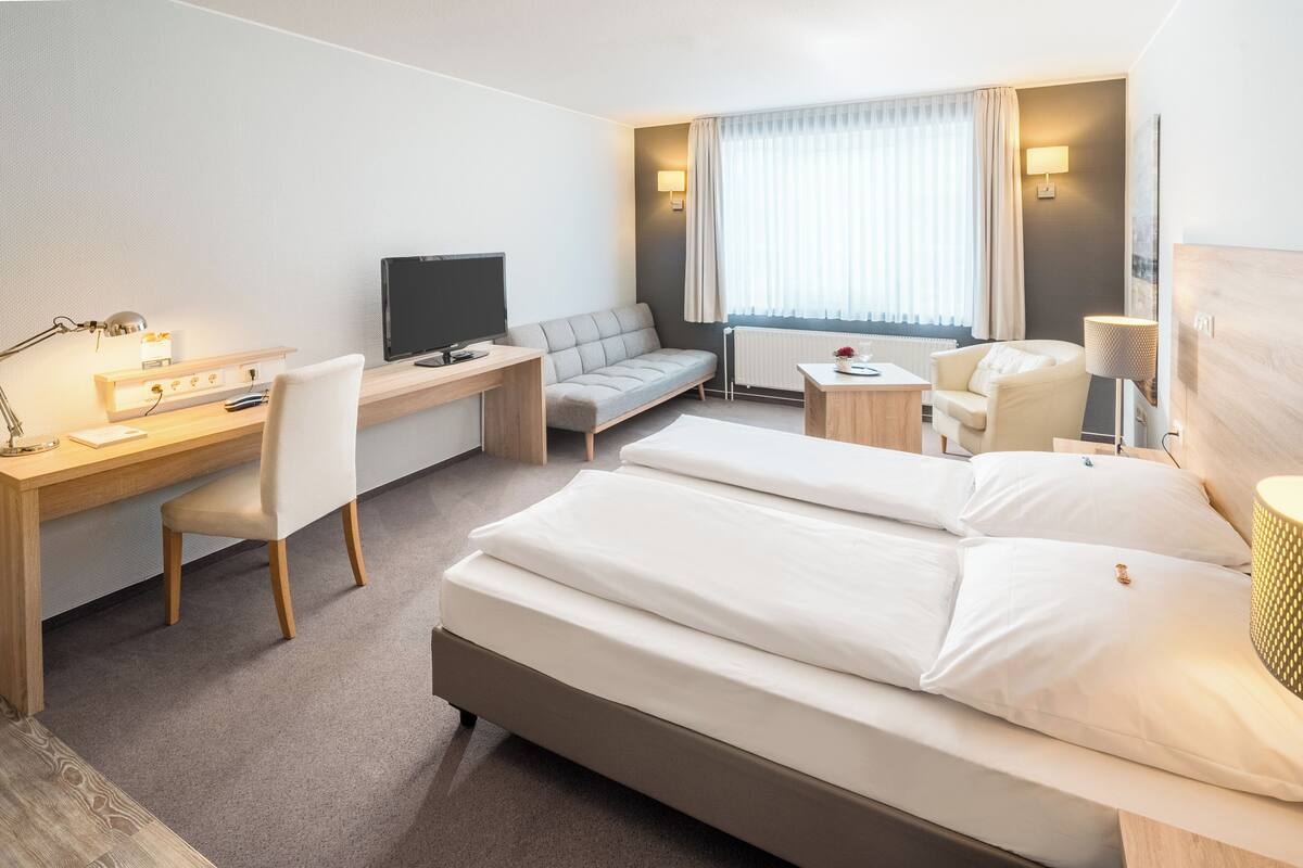A hotel room with a bed, a desk with a computer, a TV, a sofa, and a chair.