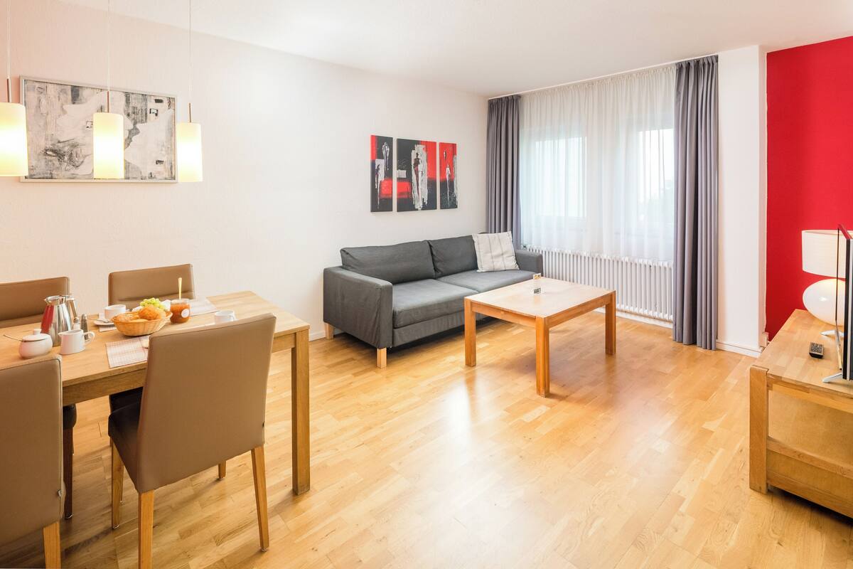 A modern living room with a grey sofa, wooden coffee table, and a dining area with chairs and a table set for breakfast.