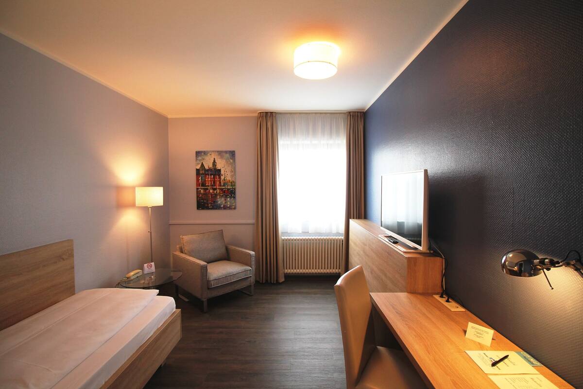 A hotel room with a bed, a desk, a chair, a TV, a lamp, and a painting on the wall.