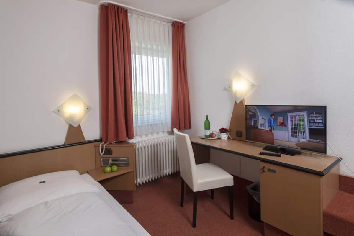A hotel room with a bed, a desk with a chair, a television, and a window with curtains.