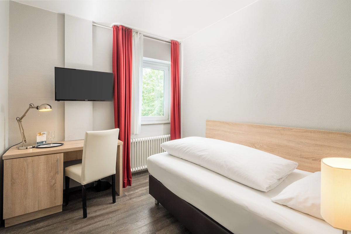 A hotel room with a bed, desk, chair, TV, window with curtains, and a lamp.