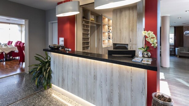 A modern reception area with a wooden reception desk, pendant lighting, and a view of a dining area with red chairs. - Hotel Westerfeld (Hemmingen)