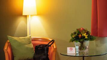 Een hotelkamer met een enkele oranje stoel, een zwarte handtas, een groen kussen, een glazen tafel met een bloevas en een lamp.