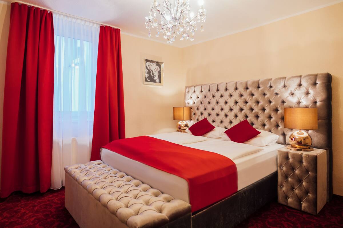 A hotel room with a large bed, red curtains, a chandelier, and a framed picture on the wall.