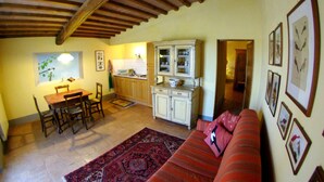 A cozy living room with a red sofa, wooden dining table, and chairs. There is a kitchen area with cabinets and a window. - Agriturismo Fattoria Santa Vittoria (Foiano della Chiana)
