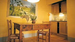 A dining area with a wooden table and chairs, a painting of a landscape, and a kitchen in the background. - Agriturismo Fattoria Santa Vittoria (Foiano della Chiana)