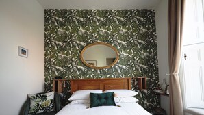 A neatly made bed with a wooden headboard, a round mirror above it, and a tropical leaf-patterned wallpaper. - The Cowrie Guest House (Berwick-upon-Tweed)