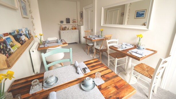 A dining area with wooden tables and chairs, a kitchen in the background, and a mirror on the wall. - The Cowrie Guest House (Berwick-upon-Tweed)