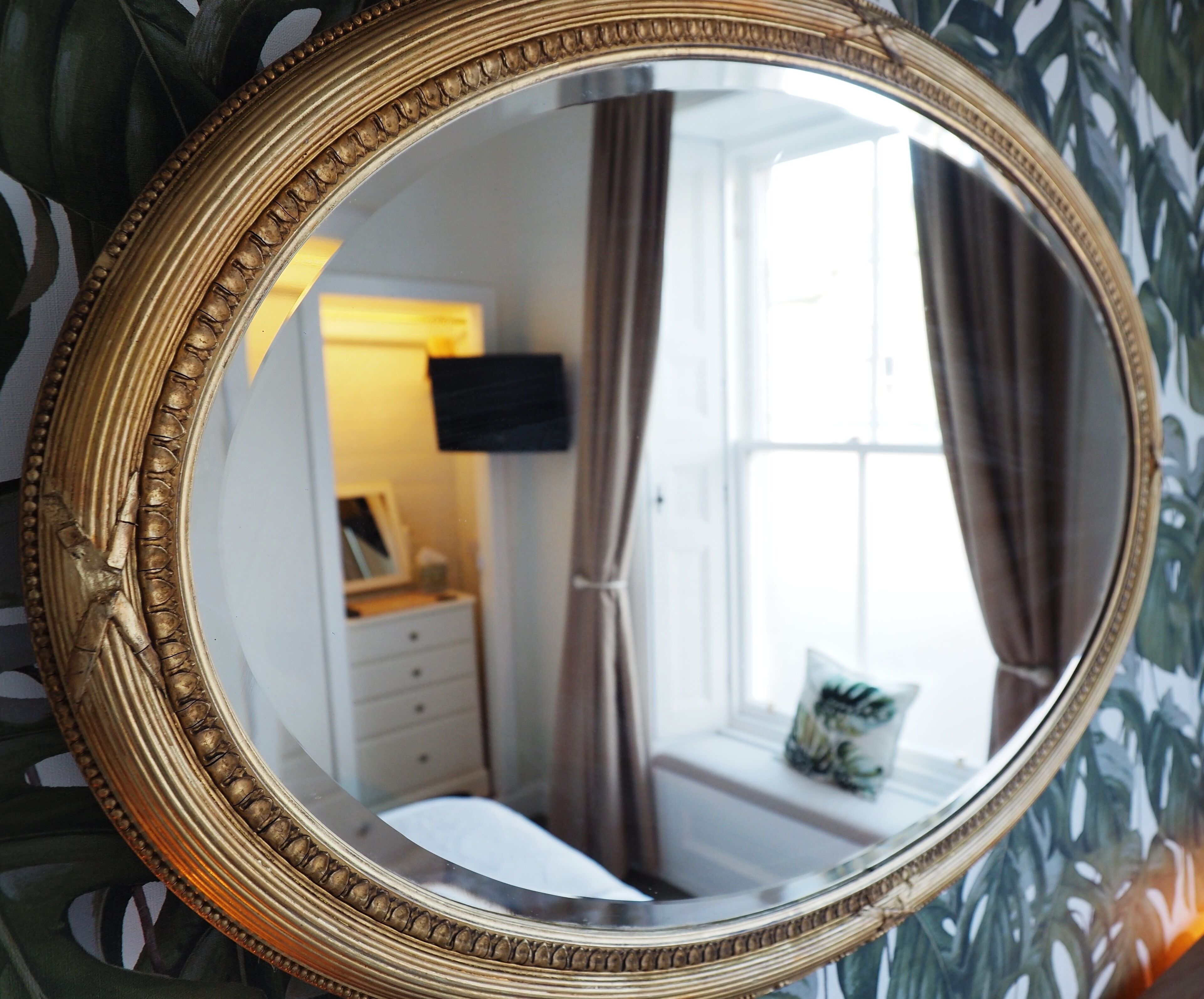 A round mirror with a decorative frame reflecting a room with a bed, a nightstand, a wall-mounted TV, and a window with curtains.