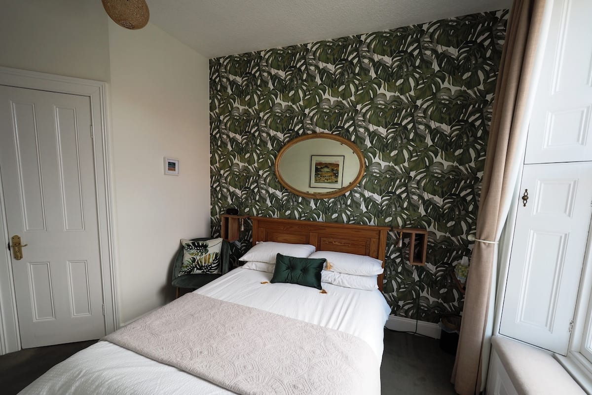 A bedroom with a bed, a wooden headboard, a wall with a tropical leaf pattern, a round mirror, and a door.