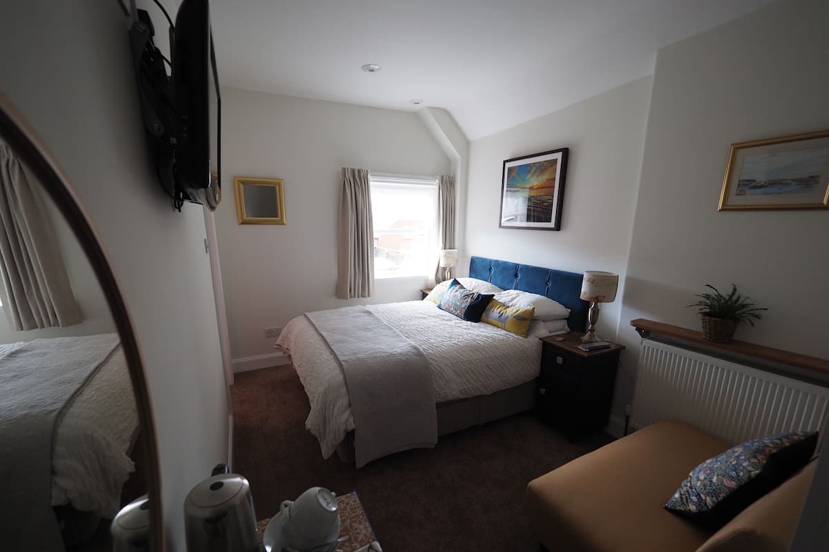 A hotel room with a bed, a sofa, a bedside table with a lamp, a window with curtains, and a radiator.