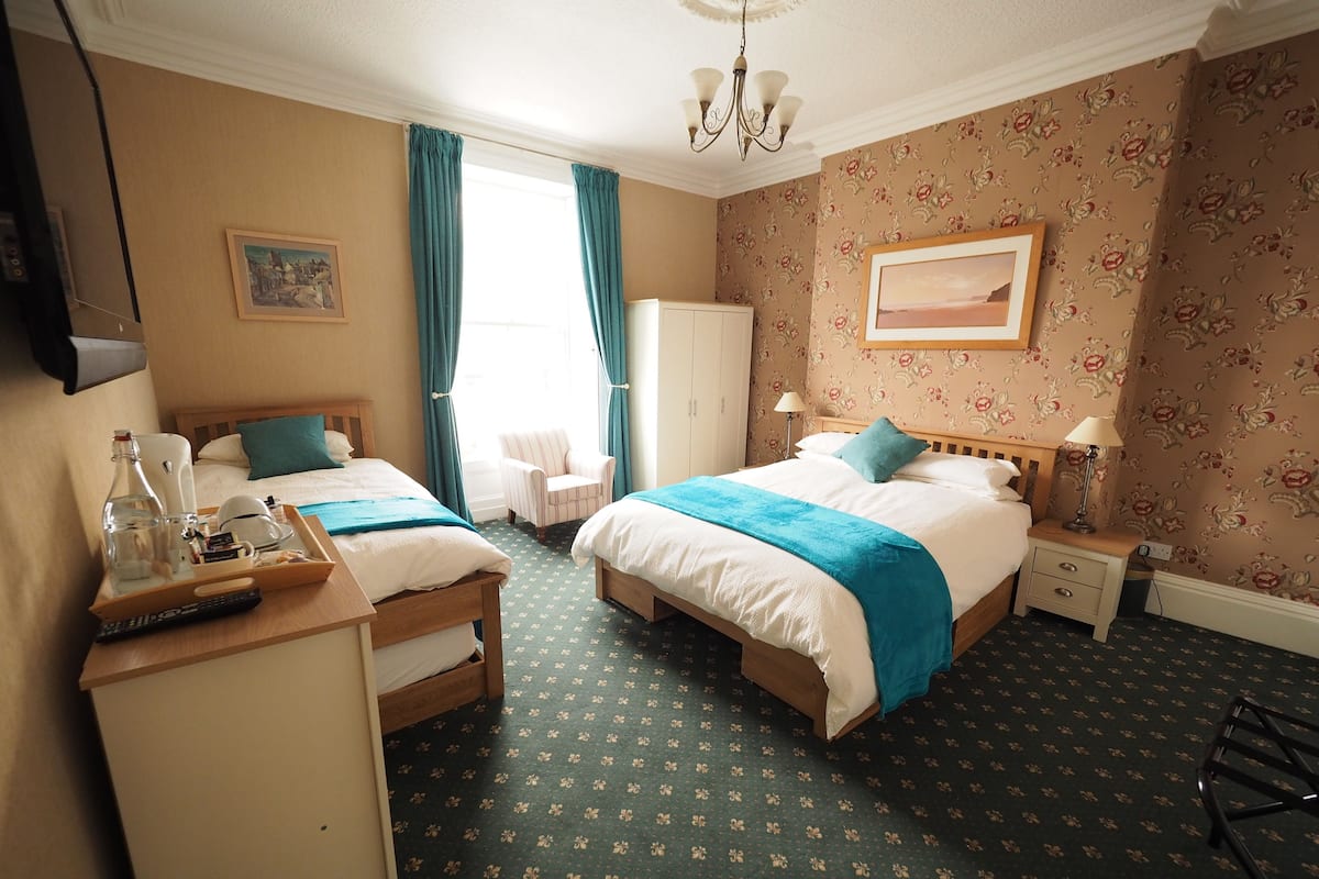 A hotel room with two beds, a desk with a tray of toiletries, a chair, a wardrobe, and a painting on the wall.