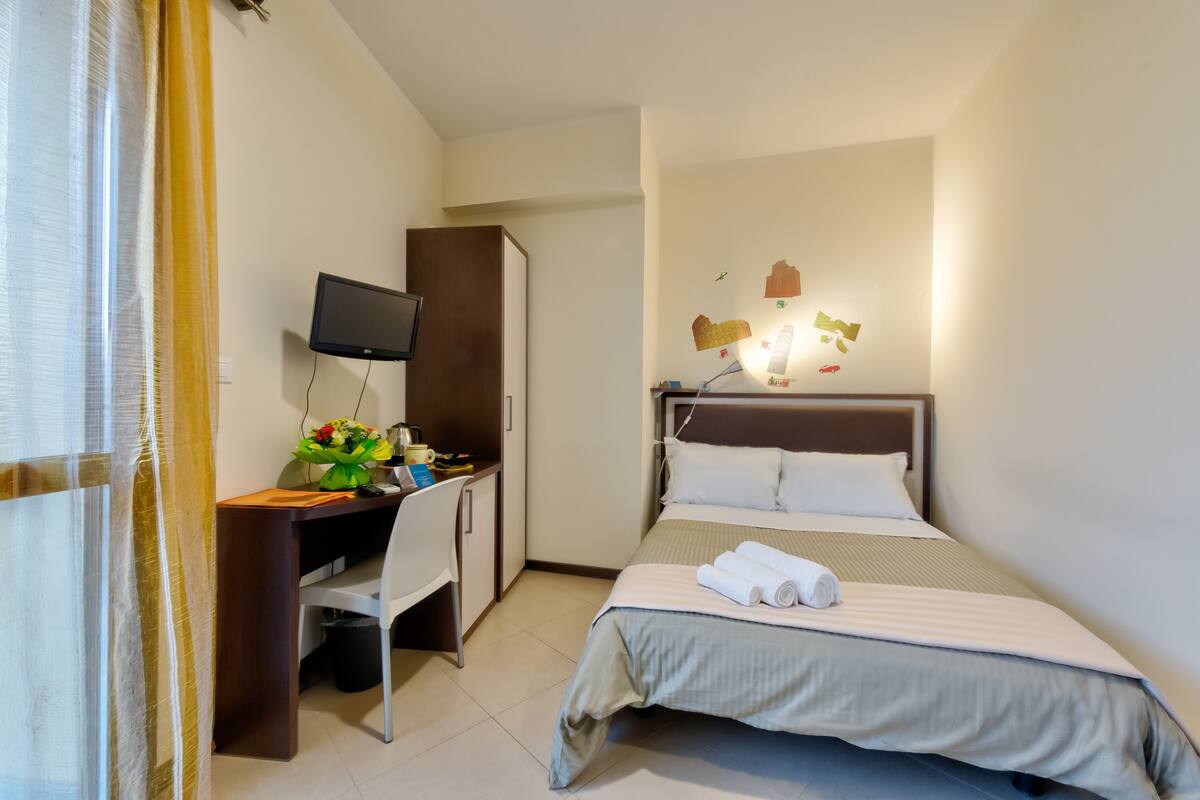 A hotel room with a bed, a desk with a chair, a TV, and a wall-mounted lamp.