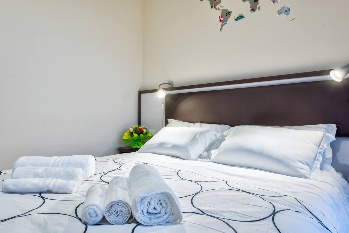A neatly made bed with white linens and rolled towels, a nightstand with a lamp, and a wall map.