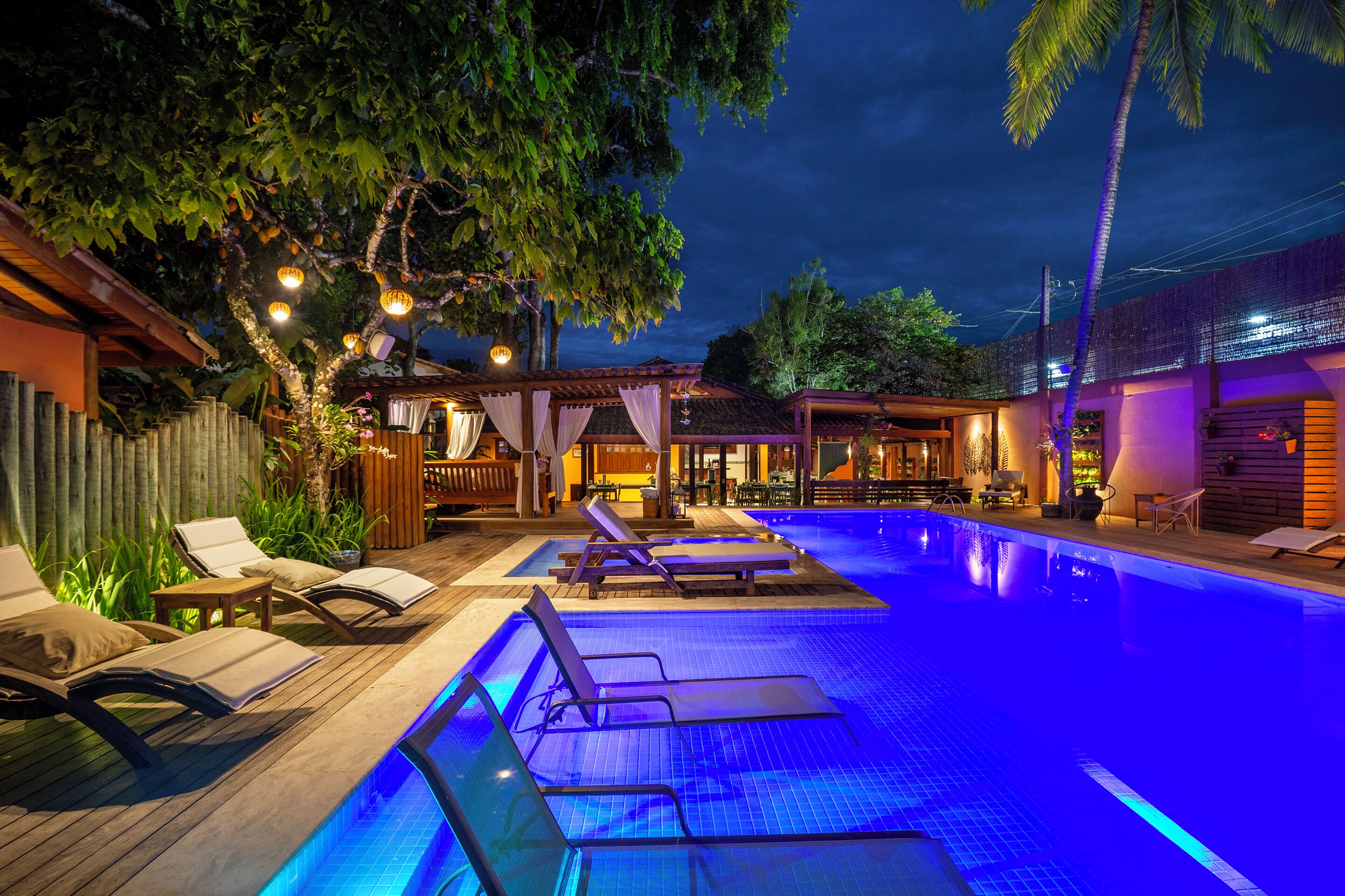 An outdoor swimming pool with lounge chairs and a wooden deck, surrounded by trees and a building with a tiled roof.