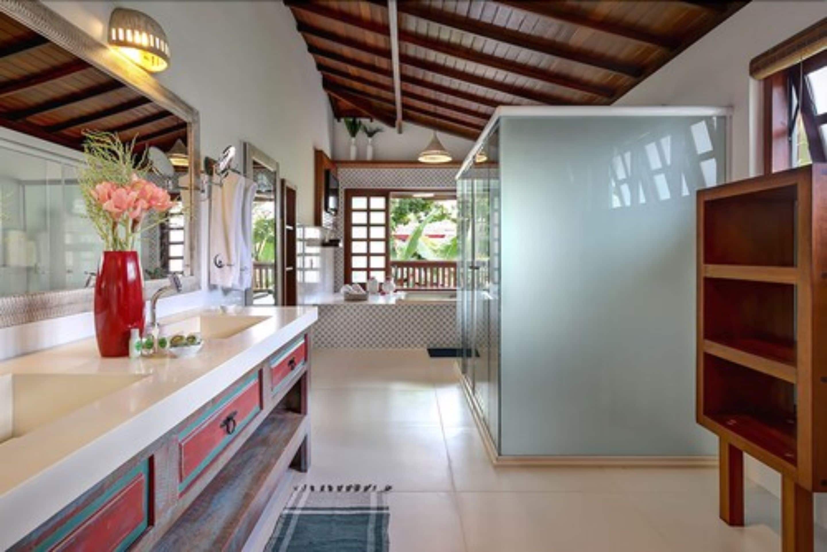 A modern bathroom with a large mirror, a red vase with flowers, a wooden cabinet, and a view of the outdoors through a window.