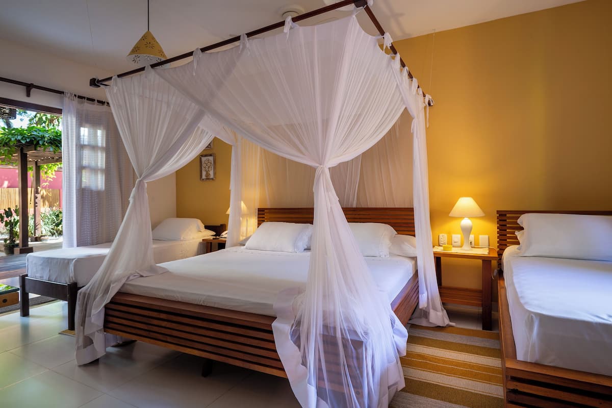 A room with a four-poster bed, two single beds, a bedside table with a lamp, and a view of greenery outside.