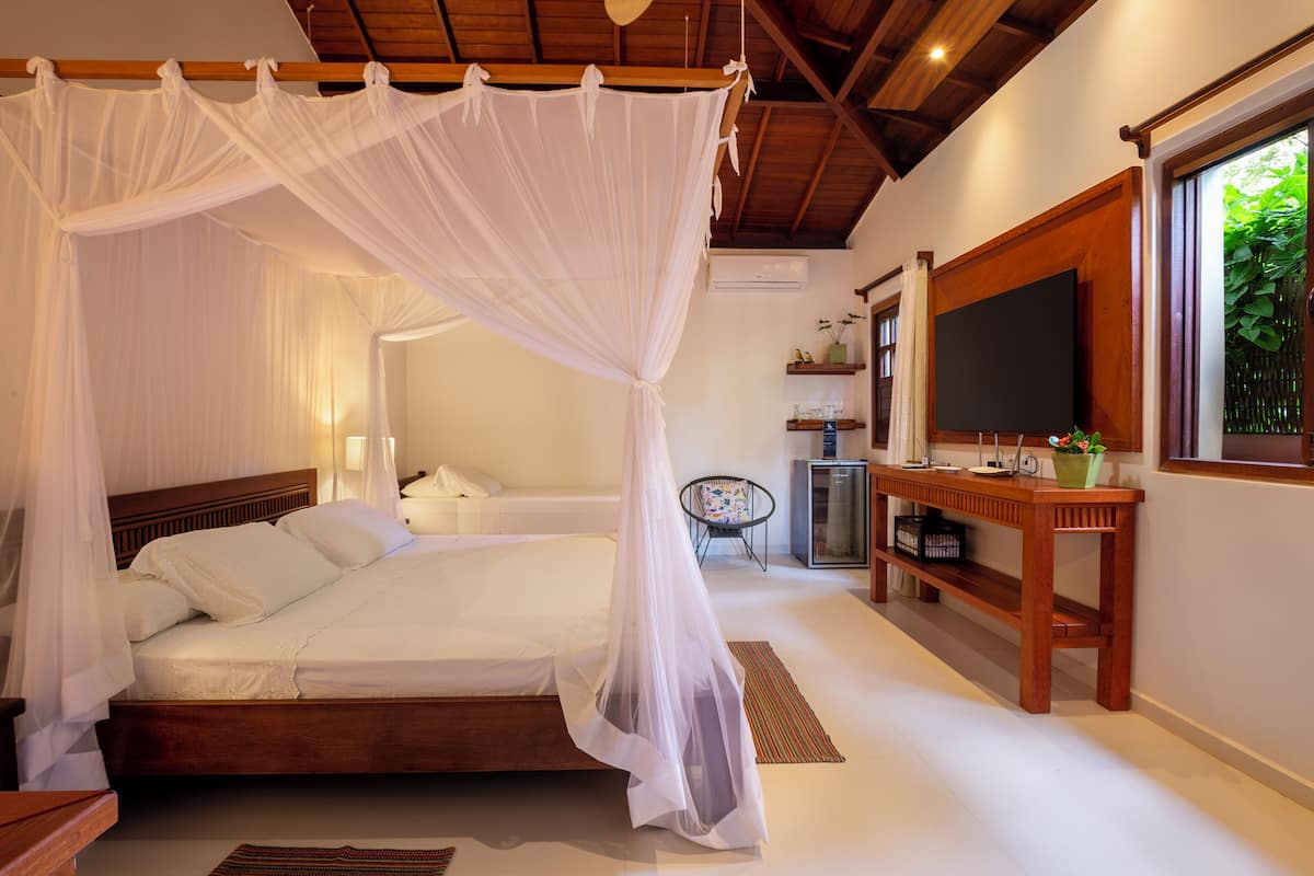 A bedroom with a canopy bed, a wooden headboard, a bedside table with a lamp, and a large window with a view of greenery.