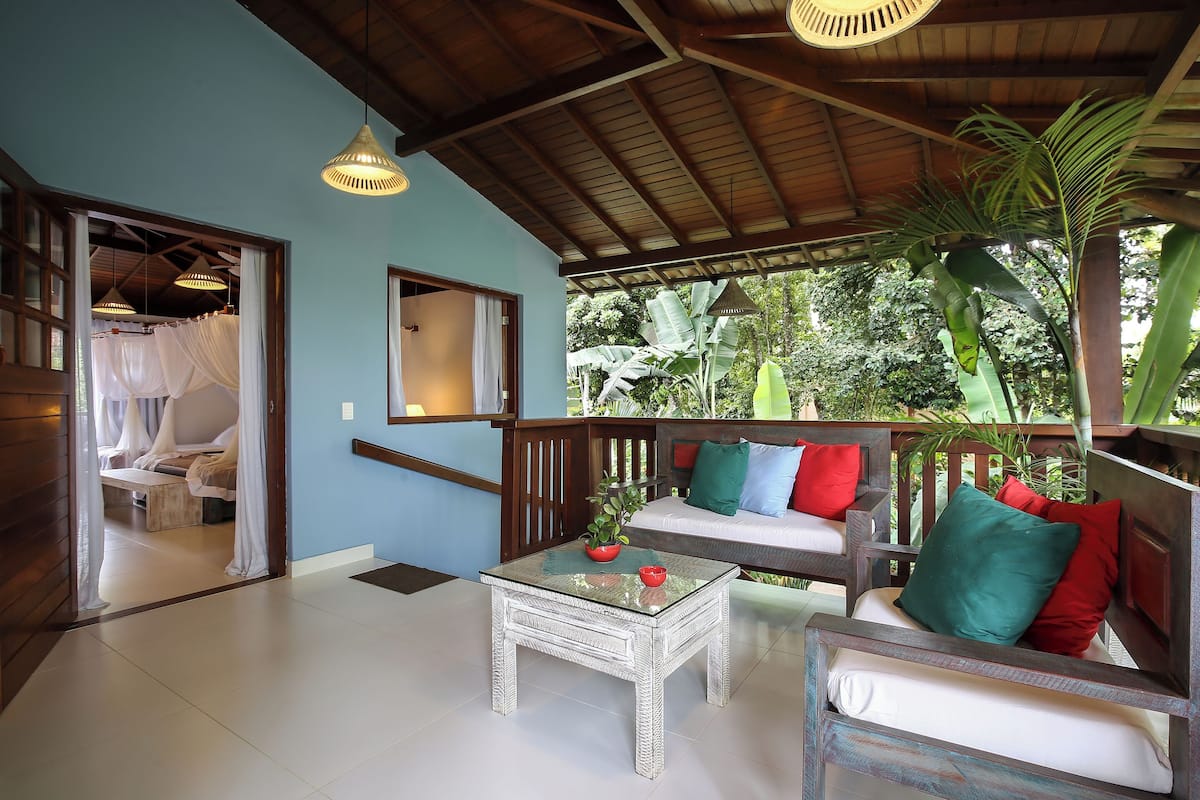 A room with a glass coffee table, a wooden sofa with cushions, a wooden bench, and a view of greenery outside.