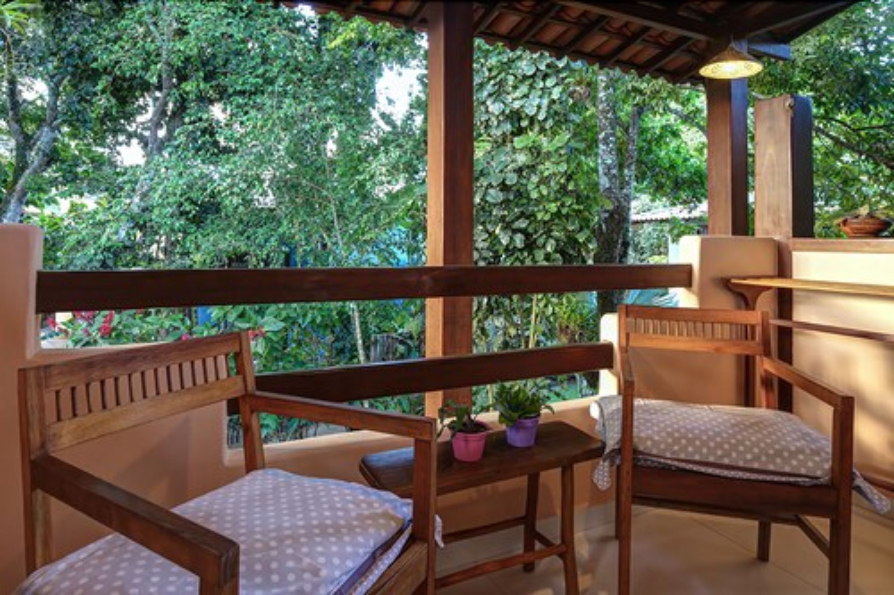 A wooden balcony with two chairs and a small table, surrounded by lush greenery.