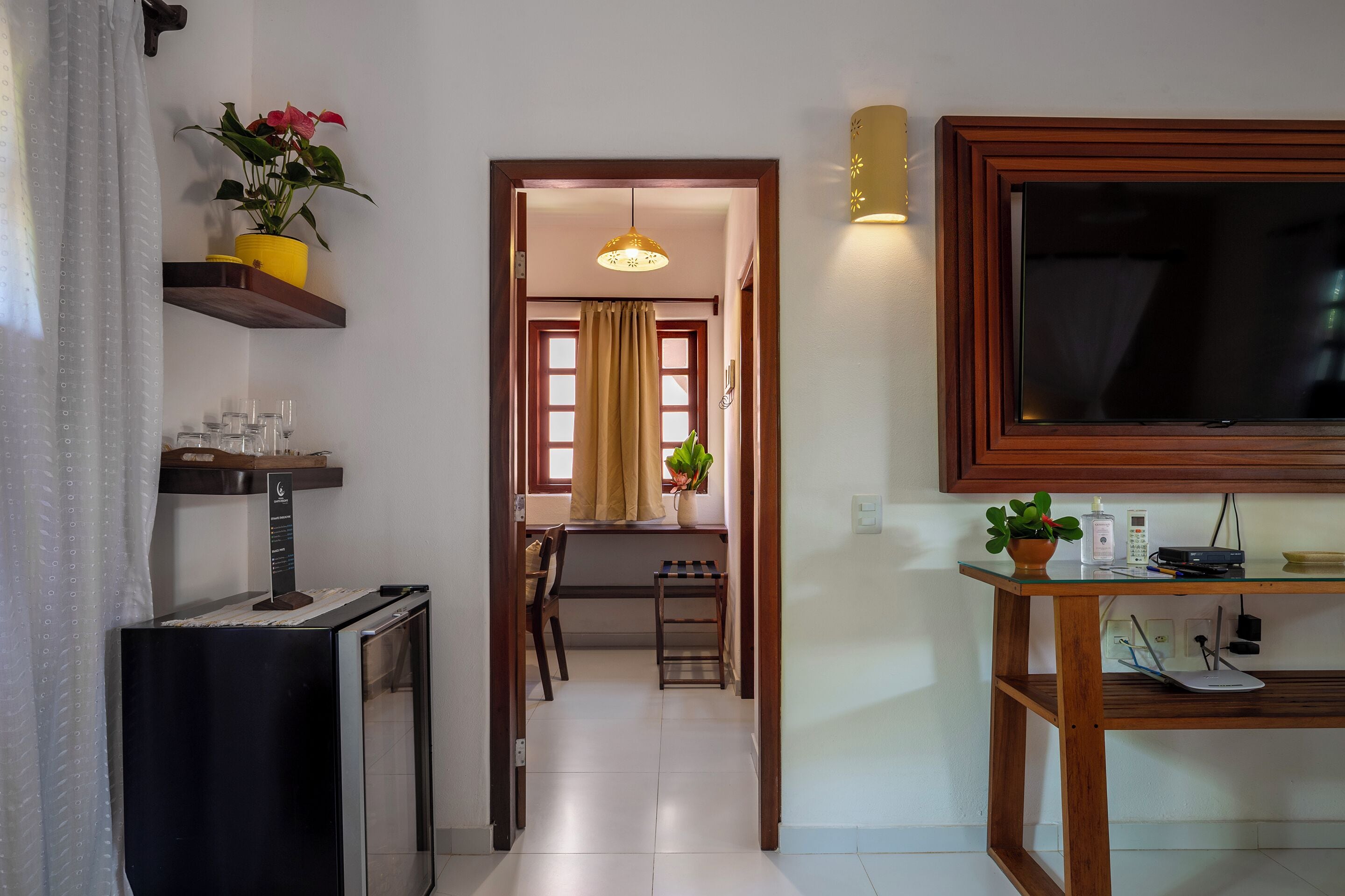 A hotel room with a TV, a small table with a plant, a shelf with decorative items, and a door leading to a dining area.
