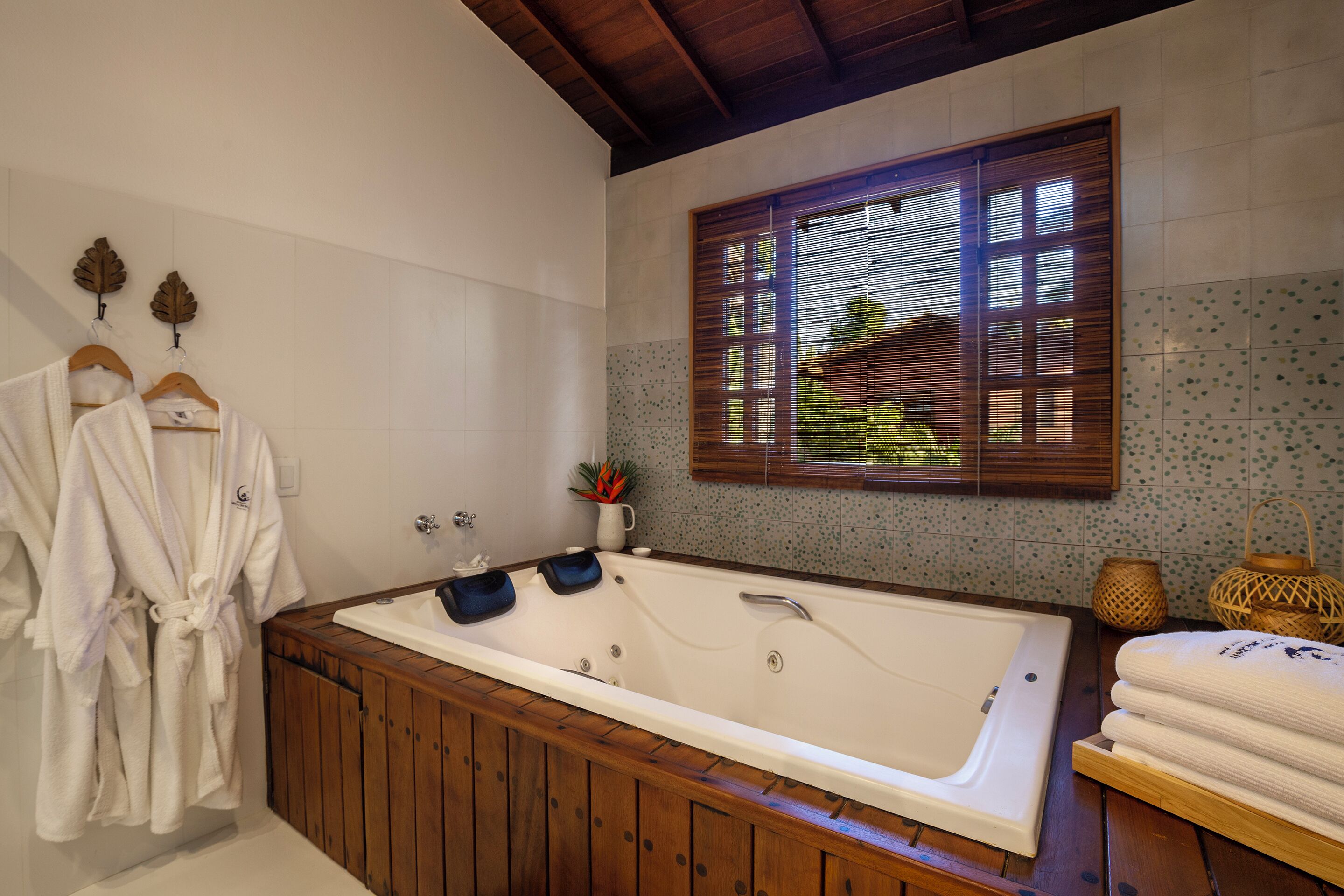 A bathroom with a wooden bathtub, white towels, and a window with blinds.